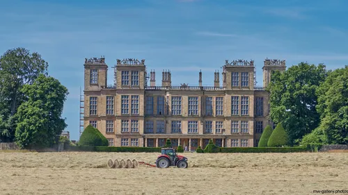 This landscape photograph captured in the late morning during the summer features Hardwick Hall, an Elizabethan manor house located in Derbyshire, United Kingdom. The imposing architecture of Hardwick Hall, a well-known National Trust property, dominates the scene with its extensive windows and ornate rooftop details. In the foreground, a red tractor is positioned on the hay-strewn grounds, highlighting rural life in the English countryside. Trees frame the stately manor, reflecting the verdant environment typical of summer. The image showcases the historic and architectural significance of Hardwick Hall, an iconic example of Elizabethan design and one of the most important manor houses in Derbyshire.